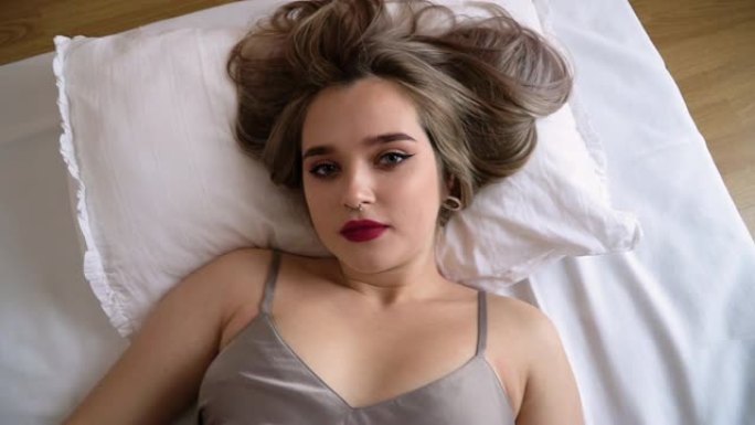年轻女士的肖像，妆容明亮，鼻子刺穿躺在床上。女人很害羞，用头毯子盖住自己。