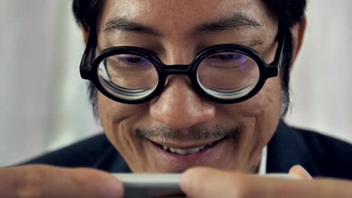 特写亚洲大胡子男人用手机带着微笑的脸，4k慢动作的亚洲男人戴着眼镜在手机上观看。