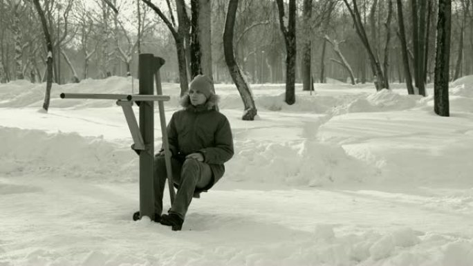 穿着蓝色羽绒服的男子在冬季公园的模拟器上做腿部锻炼。