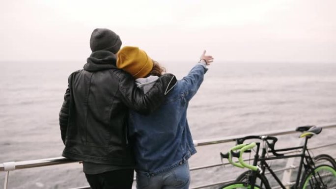 年轻男子和一个女孩站在海上木制码头上，年轻夫妇享受时光。戴着黄色帽子的女孩指着大海的地平线。秋。罕见