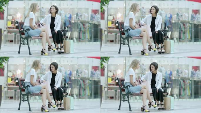 两个漂亮的年轻女子坐在购物中心的长凳上，在一起购物成功后聊天，购物袋附近有购物物品