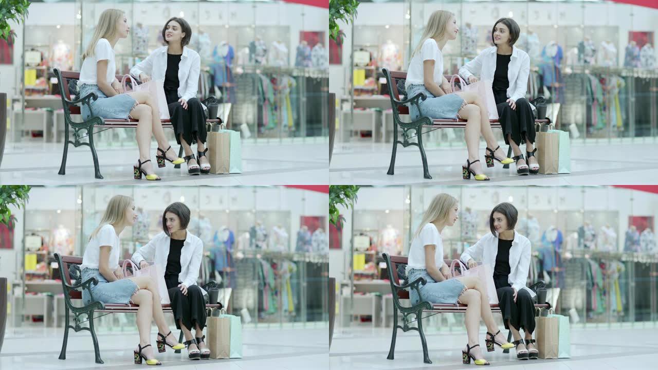 两个漂亮的年轻女子坐在购物中心的长凳上，在一起购物成功后聊天，购物袋附近有购物物品