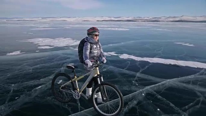 女人正走在冰上的自行车旁边。这个女孩穿着银色羽绒服，背包和头盔。冰冻的贝加尔湖的冰。自行车上的轮胎上