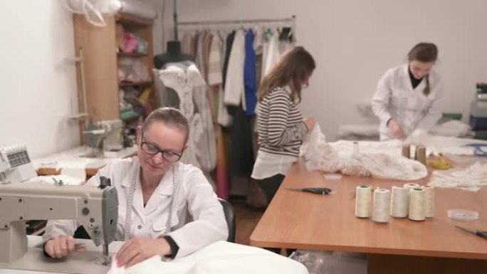 工作室的工作过程。左边的女裁缝正在缝纫机上缝制白色织物。两名裁缝站在她身后的桌子旁，正在做婚纱。