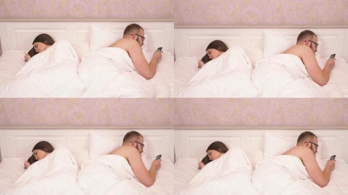 一个迷人的年轻黑发和一个戴眼镜的大胡子男人背靠背躺在床上。她正在睡觉，他拿着手机滚动