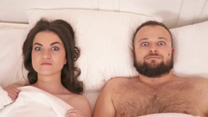 一张被带走的床上白色毯子的俯视图，一个漂亮的黑发女人，一个大胡子的男人躺在床上，瞪着眼睛，凝视着他们