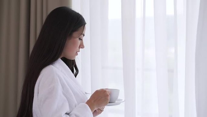一个年轻的黑发女人在窗边喝茶或咖啡的特写镜头。微风移动窗帘。她喝了一口，品尝了一下，弯下头，叹了口气