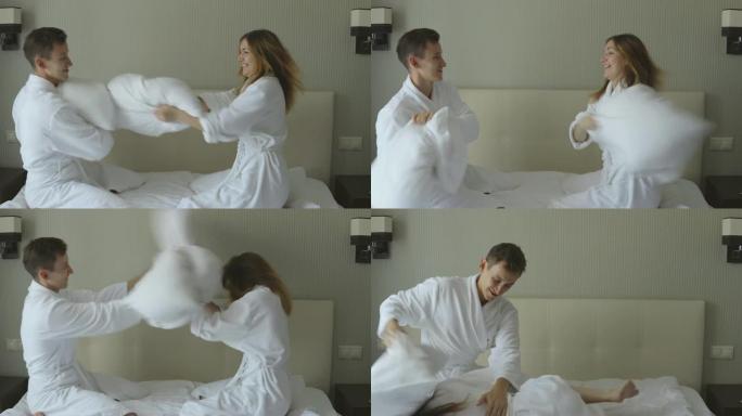 蜜月期间，可爱的年轻夫妇穿着白大褂在他的房间里傻子，在床上用枕头安排打架