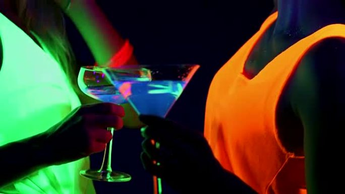 女人用UV面漆，发光的手镯，饮料，发光的衣服在镜头前一起跳舞，饮料的特写镜头。高加索人和亚洲女人。。