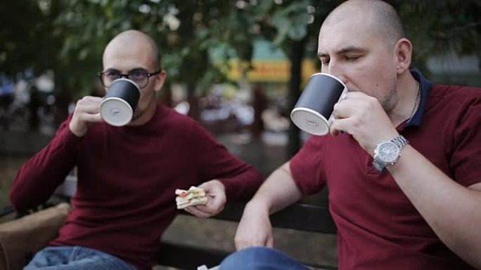 两个男人在公园的长凳上吃午餐，吃三明治和杯子里的饮料