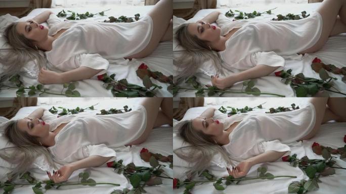 穿着短透明睡衣的年轻女士躺在床上，妆容明亮。红玫瑰在靠近女性身体的床上。慢动作。