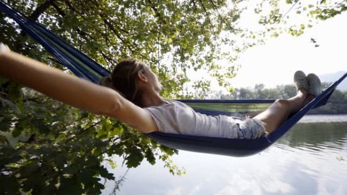 年轻女子在河边的吊床上放松，双手放在脑后享受宁静的绿色环境。人们在度假中旅行放松概念