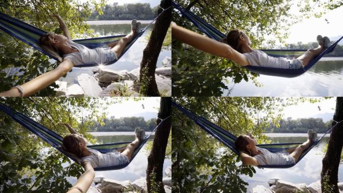 年轻女子在河边的吊床上放松，双手放在脑后享受宁静的绿色环境。人们在度假中旅行放松概念