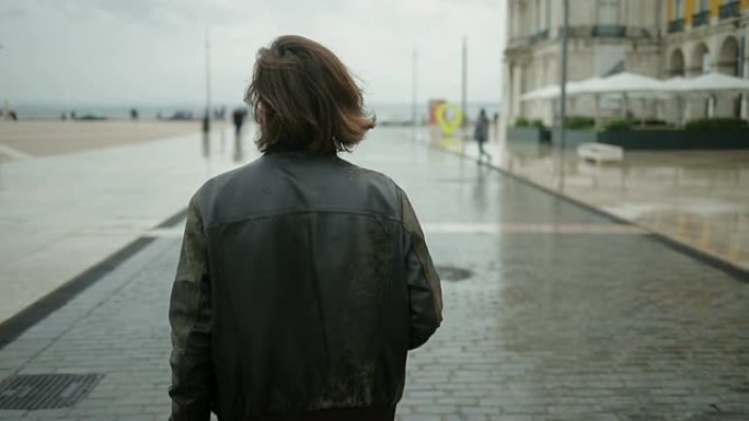 穿着旧皮夹克走在街上的年轻黑发男子的后视图。