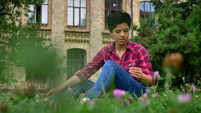 迷人的年轻大学生黑色短发坐在大学附近公园的草地上，快乐开朗