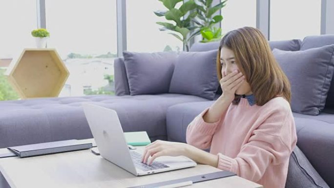 亚洲女性自由职业者在笔记本电脑上打哈欠，文书工作坐在一层楼，靠在现代公寓沙发上，在家工作的概念