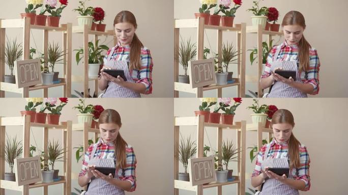 小企业。一个穿着格子衬衫和条纹围裙的女孩站着，手里拿着数字平板电脑，并计划着。花店在柜台上的花盆里做