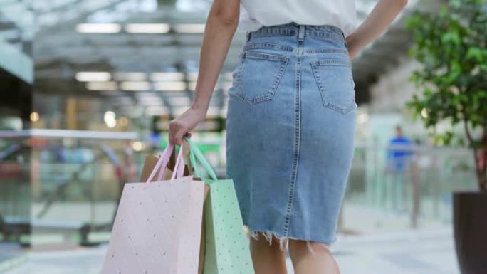 穿着牛仔铅笔裙的优雅年轻女子在购物中心走向自动扶梯的后景，在购物成功后手里拿着多色的包