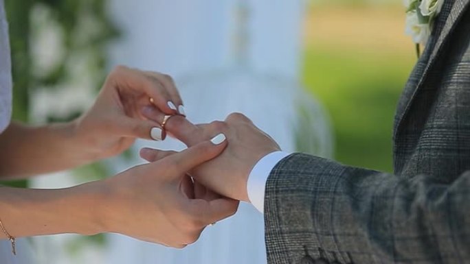 新娘和新郎在绿色自然的背景下交换结婚戒指。新郎把戒指放在新娘的手臂上特写镜头。婚礼当天。