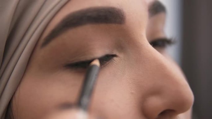 一位年轻的穆斯林妇女用黑色眼罩在眼睑上画了一个箭的特写镜头。穿着米色头巾的穆斯林妇女的惊人外观。侧视
