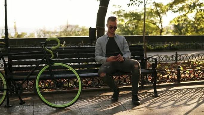 穿着蓝色衬衫的年轻人坐在长凳上，手里拿着手机发短信。骑手在公园骑徒步自行车后休息。阳光普照，绿树在背