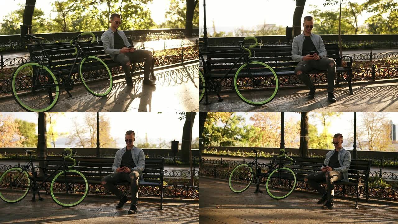 穿着蓝色衬衫的年轻人坐在长凳上，手里拿着手机发短信。骑手在公园骑徒步自行车后休息。阳光普照，绿树在背