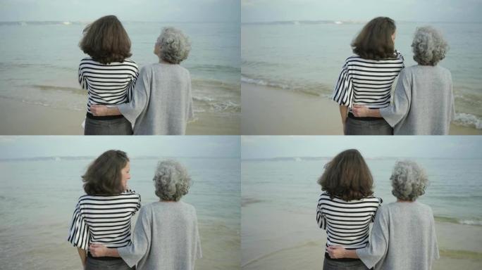 快乐的高级女人在海边拥抱高黑发女人的后视图。