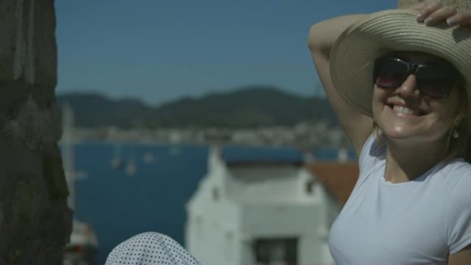 侧视图-戴着帽子的年轻女孩欣赏着大海和自然的景色，坐在石墙附近，在阳光明媚的夏日晒日光浴。