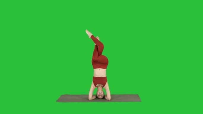 美丽的年轻女子做瑜伽运动的变化支持的倒立，鹰航salamba sirsasana，交叉腿在绿色屏幕上