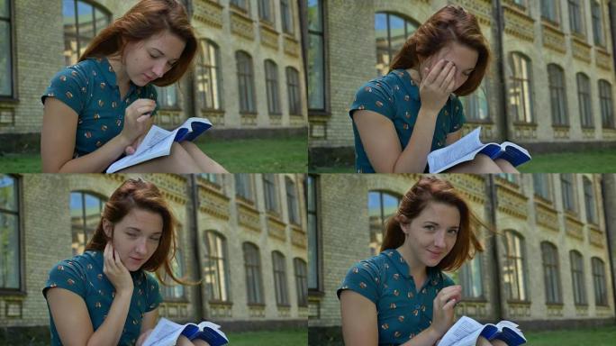 可爱的姜学生看书，对着镜头微笑，坐在大学附近的公园里，背景建筑，快乐的女性
