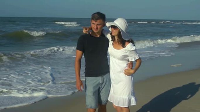 年轻美丽的夫妇在海边散步。男孩和女孩。他们很高兴。阳光灿烂。慢动作。他们站着看着远处。海浪袭击了海岸