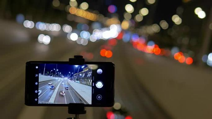 智能手机安装在三脚架上。他们在美丽的灯光背景下拍摄视频，汽车交通。晚上。