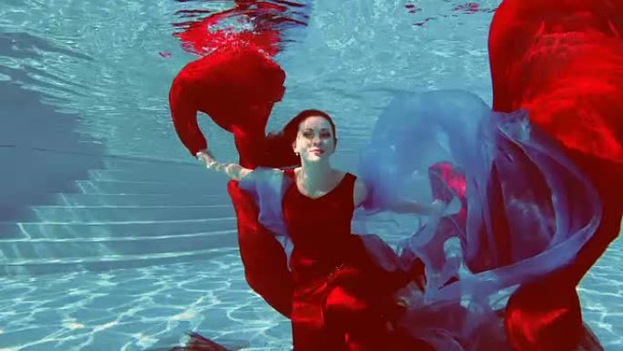 一个穿着红色连衣裙，一头红头发的美丽女孩在室外游泳池游泳并摆好姿势，手里拿着红蓝两色的布。她看着镜头