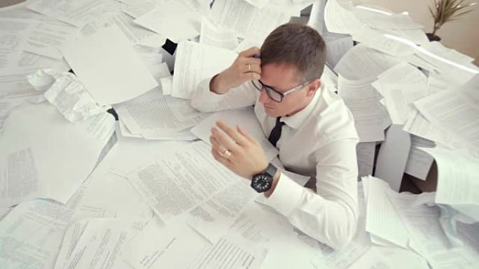 一个戴着眼镜的年轻疲倦的男人坐在一堆文件中，抬起头。由于文件数量众多，上班族无法应付工作和烦恼