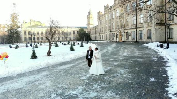 冬季婚礼。穿着婚纱的新婚夫妇正走在白雪覆盖的公园里，背景是古老的建筑和铺路石