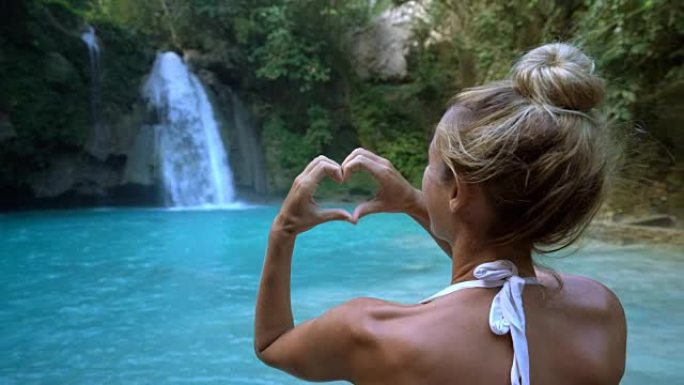 年轻女子喜欢美丽的瀑布，在菲律宾宿雾岛上制作心形手指框架。人们旅行热爱自然的概念。一个人只在和平的环
