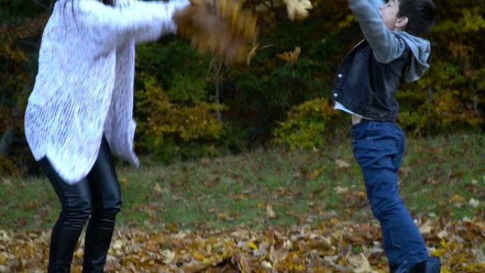 年轻美丽的母亲和她的小儿子在秋天的森林里玩得开心。他们跳起来，把树叶扔向空中。他们在笑。家庭幸福。慢