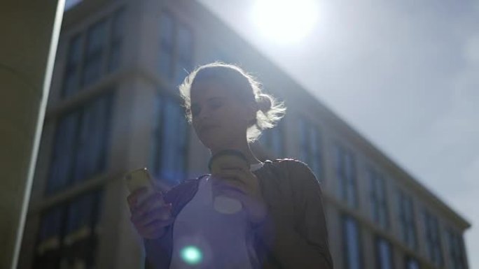 在大风天户外，年轻女子在手机上发短信的中景照片，微笑着思考该怎么回答，手里拿着外卖咖啡杯