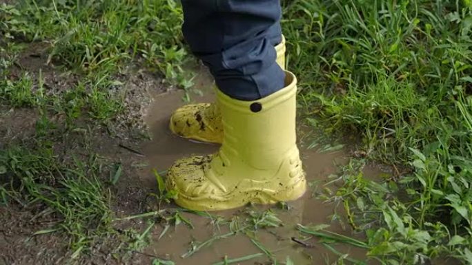 穿着亮黄色橡胶靴的小孩溅在水坑里。孩子的脚免受脏水的侵害