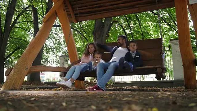快乐的年轻家庭父亲，母亲，儿子和小女儿骑着秋千在公园里。笑和微笑。4k。慢动作