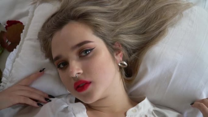 妆容明亮，鼻子穿孔的年轻女士的肖像躺在床上特写。女性慢慢地把头向右转动。