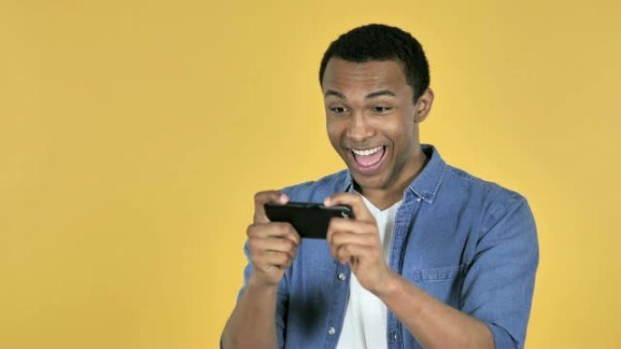 年轻的非洲男子在黄色背景上孤立的智能手机上玩游戏
