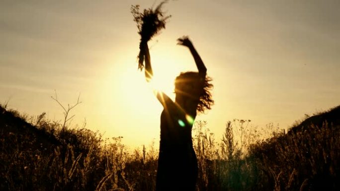一个女孩在明亮的日落背景上的黑色剪影。她在群山之间的田野里，在花丛中跳跃和挥手。底视图。