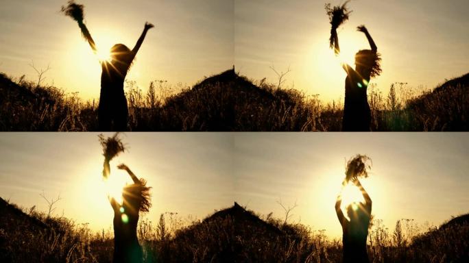 一个女孩在明亮的日落背景上的黑色剪影。她在群山之间的田野里，在花丛中跳跃和挥手。底视图。