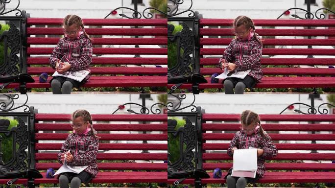 戴眼镜的女童在他的相册中画了一幅画。一个女孩坐在公园的长椅上。秋天的日子。