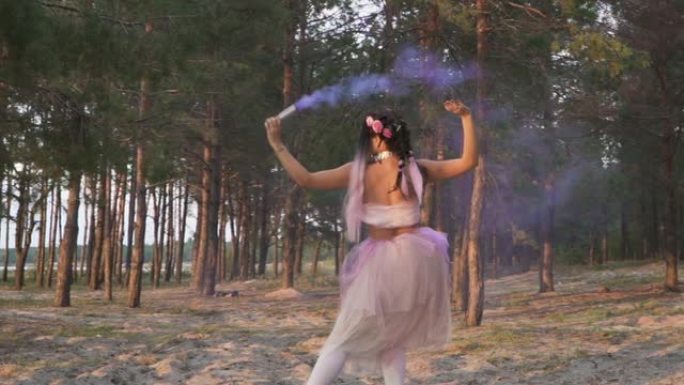 迷人的女人，穿着粉红色的连衣裙，在松树的背景上与烟雾弹共舞。一个性感女孩的舞蹈，有着美妙的发型和鲜花