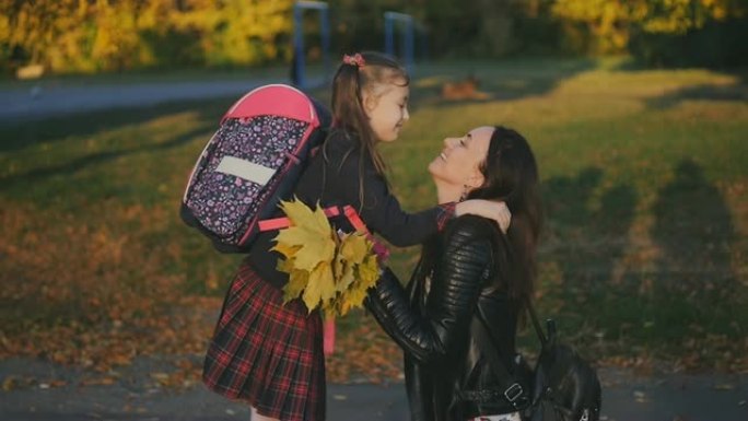 女人在学校遇见了她的小女儿。一个穿着校服，背着背包的女孩拥抱并亲吻她的妈妈。慢速射击