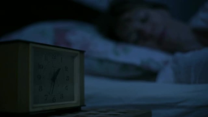 一个穿着睡衣的女孩躺在床上，看着闹钟无法入睡的特写镜头。从几个小时到面对女孩的可变焦点。美国之夜的影
