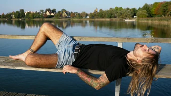 长长的金发的男模特躺在宽阔的平静湖边的码头长凳上，鸭子在湖边游泳，周围是湖边的房子和树木，阳光直射，