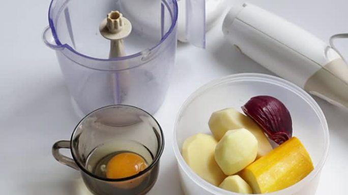 桌子上是一个用来研磨蔬菜的搅拌机。容器旁边是土豆，西葫芦和洋葱。玻璃杯里有一个鸡蛋。